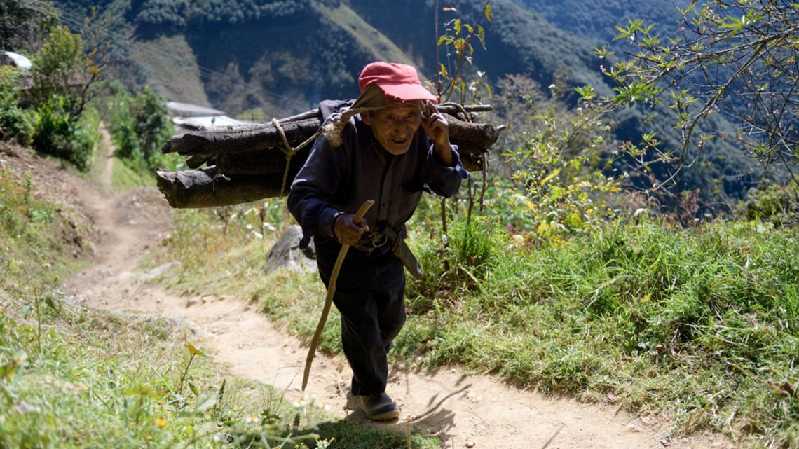 älterer Mann läuft an einem steilen Abhang in den Bergen hinauf  dabei trägt er ein Bündel Baumstämme