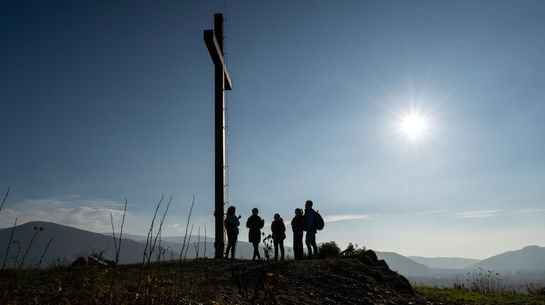 Wanderer*innen am Gipfelkreuz. Die Sonne strahlt am blauen Himmel.