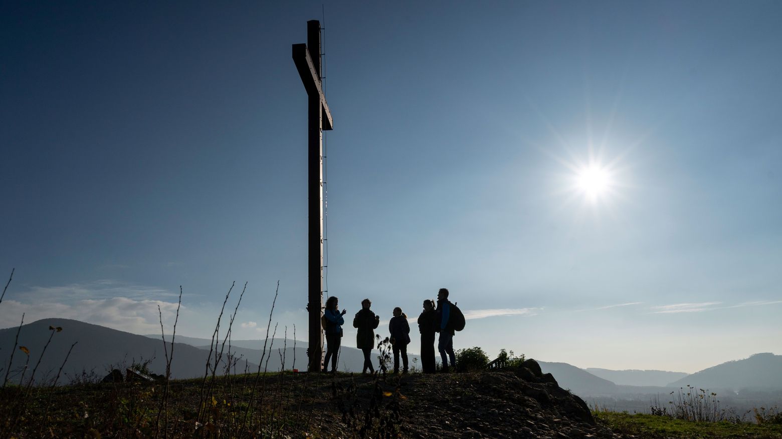 Wanderer*innen am Gipfelkreuz. Die Sonne strahlt am blauen Himmel.